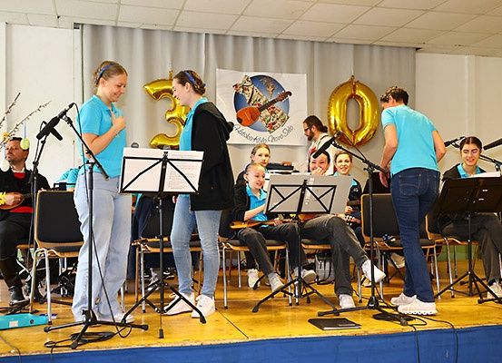 Eine Gruppe von Musikern auf der Bühne spielt Instrumente. Zwei Frauen stehen, während andere auf Stühlen sitzen. Hinter ihnen ein Banner mit einer goldenen '30' und Luftballons. Mikrofone auf Ständern.