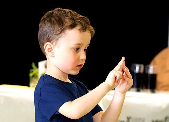 Ein junger Junge in einem blauen T-Shirt zeichnet vor einem Tisch mit zwei Gläsern dunkler Flüssigkeit. Er hat kurze Haare und konzentriert sich auf seine Aktionen.