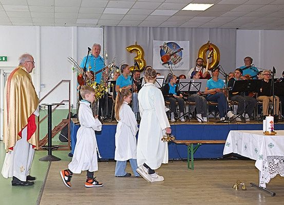Ein Priester und drei Kinder in weißen Roben gehen auf eine Bühne mit einer Band, die Instrumente spielt. Dahinter ist ein Banner mit der Nummer 30 zu sehen.