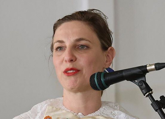 Eine Frau mit braunen Haaren, die ein weißes Spitzenkleid trägt, spricht in ein Mikrofon mit Ständer. Der Hintergrund ist eine schlichte weiße Wand.