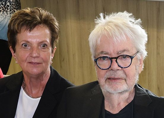 Zwei Personen, ein Mann und eine Frau, sitzen nah beieinander in einem Raum mit einer hölzernen Wand hinter ihnen.