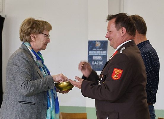 Eine ältere Frau in einem grauen Mantel und Schal überreicht einem Mann in einer braunen Uniform mit einem roten Abzeichen eine goldene Schale.