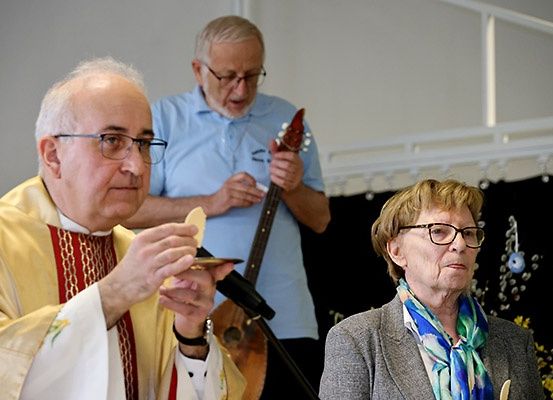 Ein Priester hält ein Stück Brot in der Hand, während ein älterer Mann eine Gitarre hinter ihm spielt und eine ältere Frau zusieht in einer Kirche.