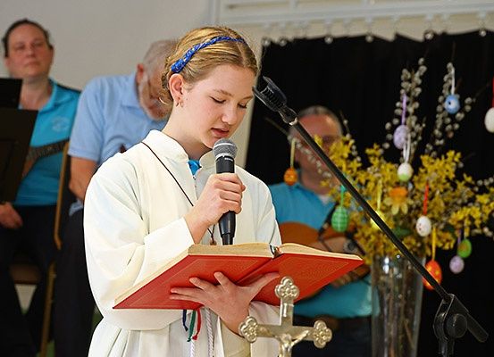 Ein junges Mädchen liest aus einem Buch und hält ein Mikrofon bei einer religiösen Veranstaltung. Hinter ihr sitzen Leute, einer spielt Gitarre. Osterdekorationen schmücken den Hintergrund.