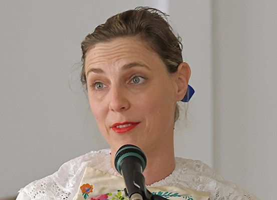 Eine Frau mit braunen Haaren spricht in ein Mikrofon und trägt ein weißes Kleid mit Blumenmustern.