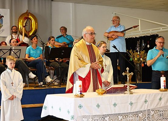 Ein Priester hält auf der Bühne mit einem Mikrofon in einer Kirche eine Predigt. Vor ihm steht ein Tisch mit einer Kerze und einem Buch. Neben ihm steht ein Kind. Hinter ihm steht eine Gruppe von Menschen, die Musikinstrumente spielen. An der Wand befindet sich ein Poster mit dem Text '0'.