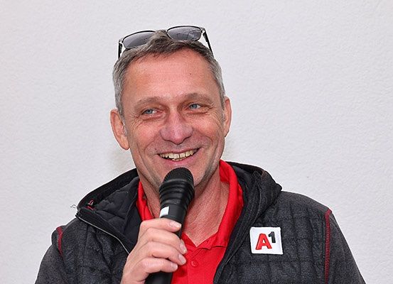 Ein Mann mit grauen Haaren lächelt, während er ein Mikrofon hält, trägt ein rotes Hemd und eine Sonnenbrille auf dem Kopf.