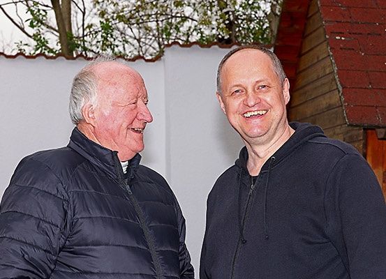 Zwei Männer, einer mit einer schwarzen Kapuzenjacke und der andere mit einer Daunenjacke, stehen vor einem Gebäude mit einem roten Dach. Sie lächeln sich an.