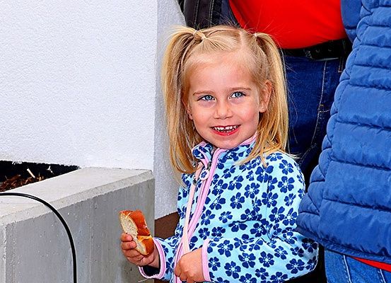 Ein kleines Mädchen in einer blauen Blumenjacke hält ein Sandwich und lächelt fröhlich, während es vor einer weißen Wand steht.