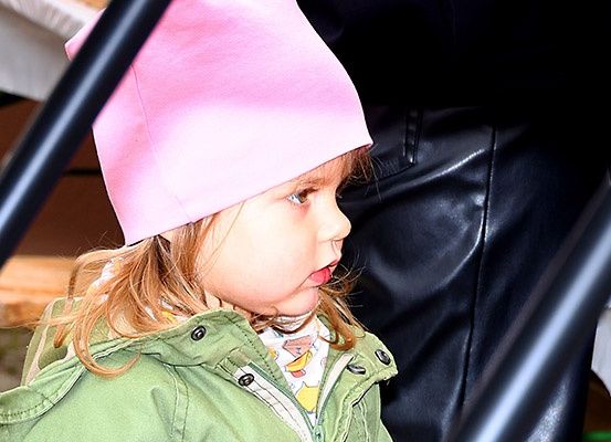 Ein kleines Mädchen mit blonden Haaren trägt einen rosa Hut und eine grüne Jacke. Es steht draußen und schaut nach unten. Ein Mensch in schwarzen Lederhosen steht hinter ihm.