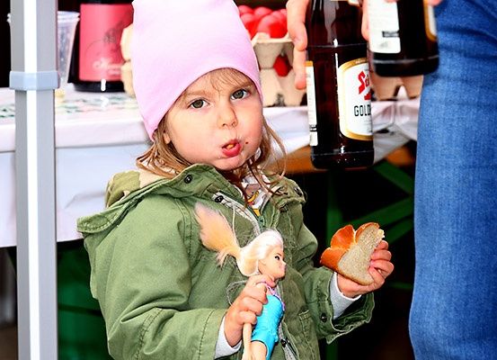 Ein junges Mädchen mit einer Puppe, isst ein Sandwich. Hinter ihr hält eine Person eine Flasche Bier. Im Hintergrund sind Flaschen und ein Tisch mit Essen.