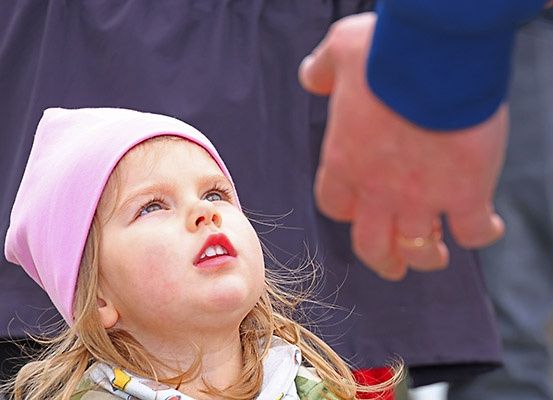 Ein kleines Mädchen in einer rosa Mütze schaut mit offenem Mund nach oben, eine Hand reicht von der rechten Seite aus.