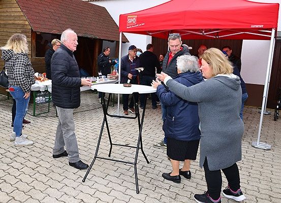 Menschen sind unter einem roten Zelt versammelt, möglicherweise für eine soziale Veranstaltung. Ein älterer Mann macht ein Foto, während ein anderer Essen serviert.