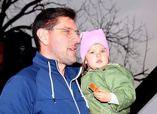 Ein Mann in einem blauen Pullover hält ein kleines Mädchen mit einer rosa Mütze und einer grünen Jacke. Sie lächeln beide, und das Mädchen hält ein Stück Orange. Der Hintergrund ist unscharf.