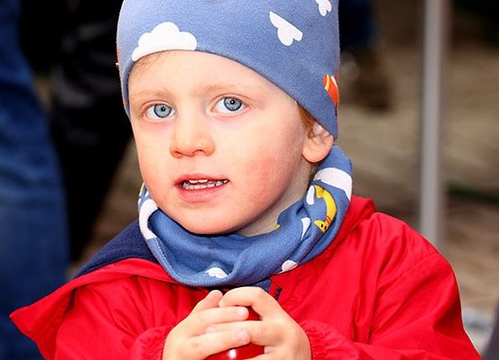 Ein junger Junge mit blauen Augen trägt einen blauen Hut und eine blaue Halsschleife und schaut nach rechts, während er ein rotes Objekt hält.