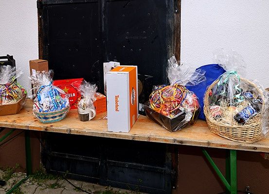 Ein Holztisch zeigt verschiedene Geschenkkörbe, darunter einer mit einer Tasse und einer weißen Box mit der Aufschrift 'Thunderbird'. Die Körbe sind in Plastik verpackt und enthalten verschiedene Artikel. Hinter dem Tisch befindet sich eine schwarze Tür mit Einschusslöchern.