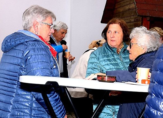 Eine Gruppe älterer Frauen und Männer steht um einen Tisch herum. Sie unterhalten sich und halten Tassen. Eine Frau mit grauen Haaren und einer Brille hält eine Tasse. Eine andere Frau in einer blauen Jacke hält eine Schachtel. Der Hintergrund ist unscharf.