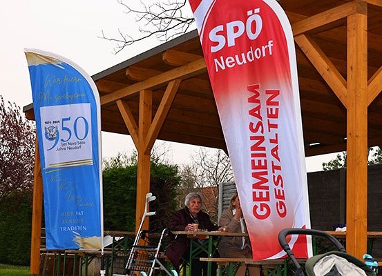 Zwei ältere Frauen sitzen an einem Tisch in einem hölzernen Pavillon, mit einem Kinderwagen in der Nähe. Das Banner hinter ihnen steht 'Spo Neudorf'. Die Frau auf der linken Seite hält einen Becher, während die andere einen Stock hält.