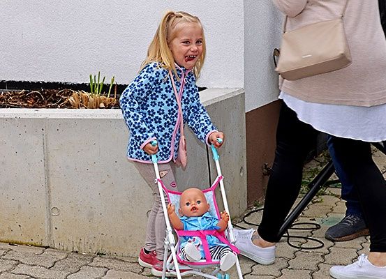 Ein kleines Mädchen weint, während es einen rosafarbenen Kinderwagen mit einer Babypuppe darin schiebt. Hinter ihr stehen zwei Erwachsene, einer mit einer beigefarbenen Handtasche. Der Hintergrund zeigt eine Betonmauer mit einer Pflanze.