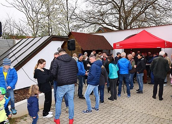 Eine Gruppe von Menschen steht in einer Schlange vor einem Gebäude mit einem roten Zelt, wahrscheinlich auf ein Ereignis wartend. Einige tragen Winterkleidung.