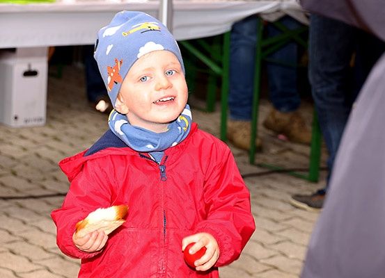 Ein junger Junge in einem roten Mantel hält ein Stück Brot und lächelt hell in einer Außenumgebung.