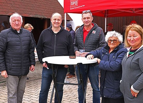 Fünf Personen stehen unter einem roten Zelt, möglicherweise bei einer Veranstaltung im Freien. Sie lächeln und scheinen für ein Foto zu posieren. Der Mann in der Mitte hält einen Becher.