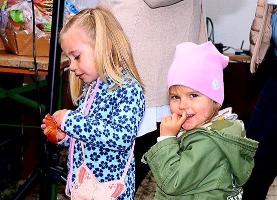 Zwei kleine Mädchen, eines in einem blauen Blumenkleid und das andere in einer rosa Mütze, stehen eng beieinander, wahrscheinlich bei einer Veranstaltung. Ein Mädchen hält einen roten Gegenstand, möglicherweise eine Süßigkeit, während das andere die Hand am Kinn hat. Hinter ihnen steht eine Person in einer beigen Jacke in der Nähe eines Schreibtisches mit verschiedenen Gegenständen.