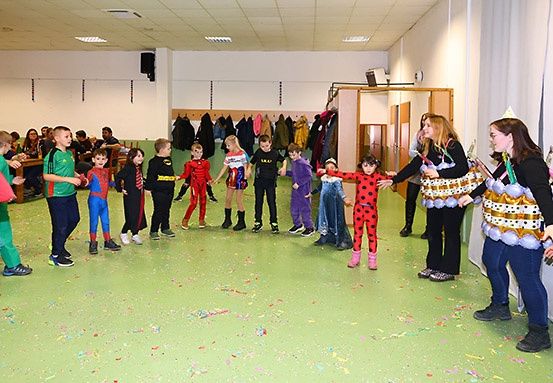Gruppe von Kindern in Kostümen tanzt mit zwei Erwachsenen auf grünem Boden mit Konfetti.