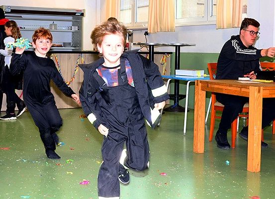Zwei Kinder in Feuerwehruniformen laufen auf einem grünen Boden mit Konfetti, während ein Mann mit Brille an einem Tisch sitzt.