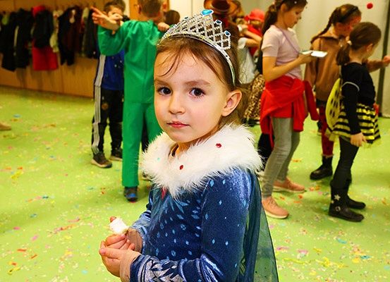 Ein kleines Mädchen, das als Prinzessin gekleidet ist, steht in einem mit Konfetti gefüllten Raum. Hinter ihr stehen andere Kinder in verschiedenen Kostümen.
