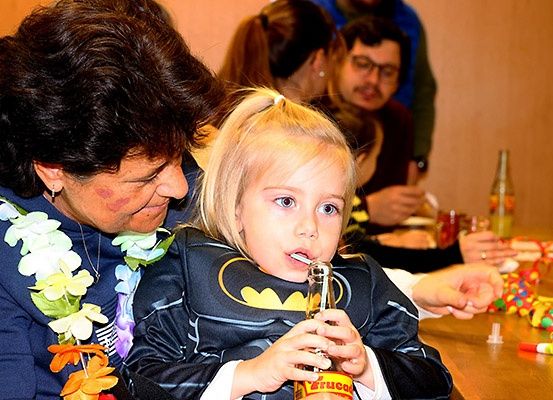 Ein kleines Mädchen in einem Batman-Kostüm trinkt aus einer Flasche, während es auf dem Schoß eines Erwachsenen sitzt bei einem festlichen Zusammentreffen. Dahinter sitzen mehrere Personen an einem Tisch mit Getränken und Spielzeug.