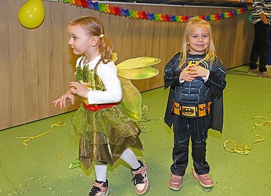 Zwei kleine Mädchen in Kostümen, eines als Fee und das andere als Batman, gehen auf einem grünen Boden mit Konfetti. Dahinter befindet sich eine Wand mit bunten Dekorationen und einem Ballon. Eine Person steht im Hintergrund.