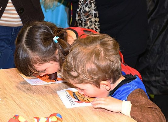 Zwei Kinder sitzen an einem Tisch, sie essen etwas, vielleicht Kuchen, und tragen blaue Jacken.