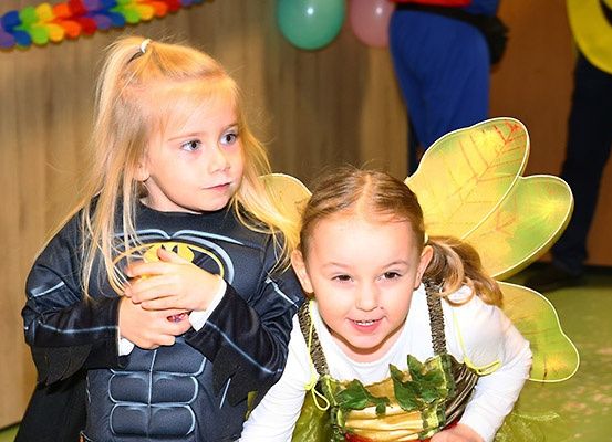 Zwei junge Mädchen in Kostümen lächeln in die Kamera. Ein Mädchen ist als Batman verkleidet, das andere als Fee. Dahinter befinden sich bunte Luftballons und eine Wand mit einer Girlande.