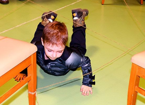 Ein junger Junge in einem schwarzen und grauen Kostüm krabbelt auf einem grünen Boden und hält an an Seile gebundenen Stühlen fest.