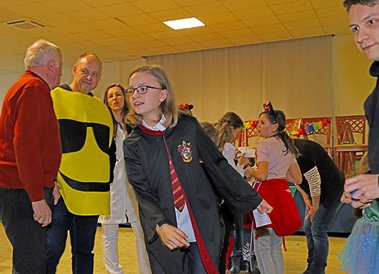 Mehrere Personen befinden sich in einem Raum, wahrscheinlich auf einer Party. Ein Mädchen in einem Harry-Potter-Kostüm steht davor. Der Raum ist mit Stühlen und Luftballons dekoriert.