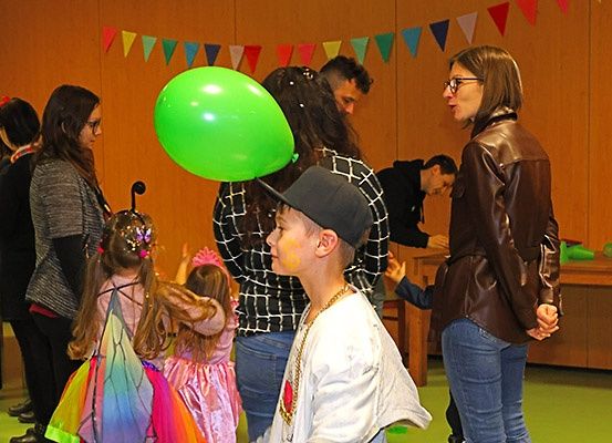 Eine Gruppe von Menschen ist in einem Raum versammelt. Einige Kinder sind in Kostümen gekleidet. Ein Junge hält einen grünen Ballon. Eine Frau steht in der Nähe und trägt eine Brille. Ein Schreibtisch ist vorhanden.