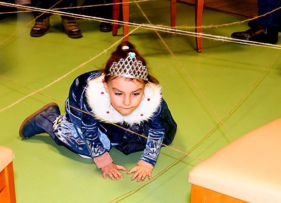 Ein junges Mädchen in einem blauen Kleid mit Krone kriecht auf einem grünen Boden, umgeben von Stühlen. Sie trägt Stiefel und einen weißen Pelzkragen. Eine Person steht in der Nähe.