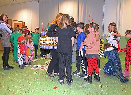 Eine Gruppe von Kindern und Erwachsenen steht in einem Raum, einige in Kostümen, mit einem konfettibedeckten Boden. Eine Frau steht in der Nähe, und ein Geburtstagskuchen ist vorhanden.