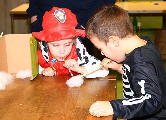 Zwei Kinder sind in einer verspielten Aktivität engagiert, eines verkleidet als Feuerwehrmann mit rotem Hut und das andere in einem Skelettkostüm. Sie pusten auf Strohhalme über einen Tisch mit Baumwolle.
