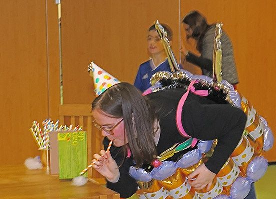 Eine Frau in einer bunten Ballon-Verkleidung und einer Party-Mütze bläst auf einen Strohhalm an einem Tisch. Ein Mädchen mit einem Pokal steht hinter ihr.