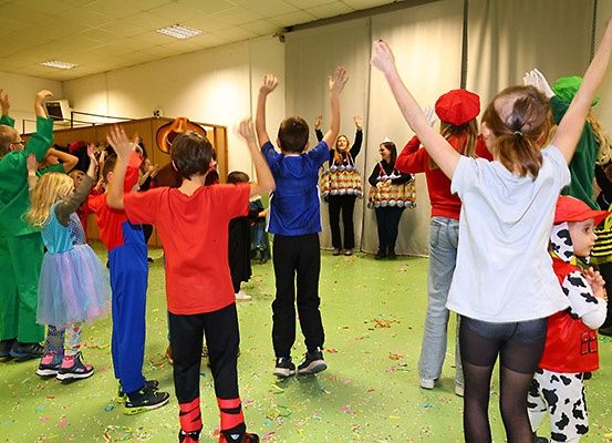 Kinder in verschiedenen Kostümen und Hüten führen einen Tanz mit erhobenen Händen in einem grün gefliesten Raum auf, wobei einige Erwachsene Trommeln im Hintergrund halten.