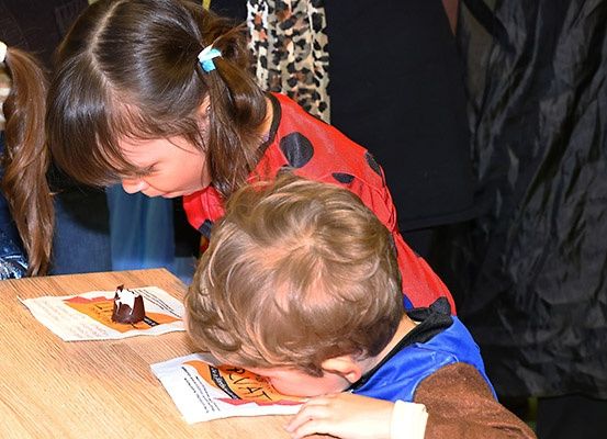 Ein Kind isst einen Schokoladenkuchen, während ein anderes Kind zuschaut. Dahinter beobachtet eine Frau in einem Punktekleid. Die Szene spielt sich drinnen ab.