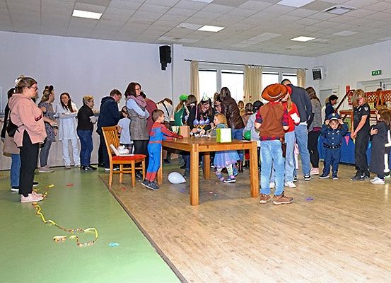 Indoor-Veranstaltung mit Erwachsenen und Kindern, einige in Kostümen, stehen um einen Tisch herum und in der Nähe einer Wand mit farbenfrohen Dekorationen.