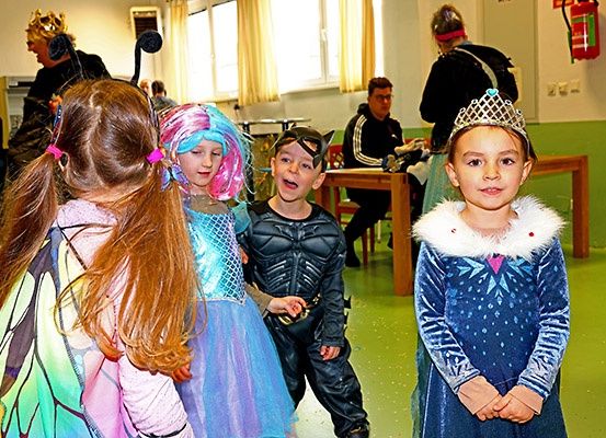 Eine Gruppe von Kindern in Kostümen steht in einem Raum. Einige sind als Prinzessinnen gekleidet, während andere als Superhelden verkleidet sind. Hinter ihnen sitzen ein Mann und eine Frau an einem Tisch.