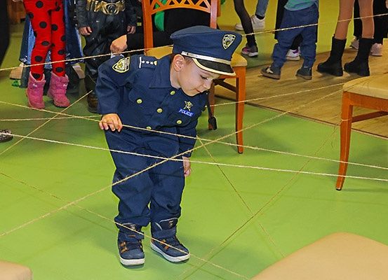 Ein junger Junge in einer Polizeiuniform läuft auf einem grünen Boden mit einem labyrinthartigen Muster aus Schnüren, umgeben von anderen Kindern.