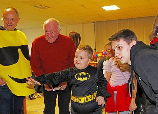 Ein Junge in einem Batman-Kostüm geht an einem älteren Mann in einem Smile-Gesichtskostüm vorbei, während ein junger Junge zuschaut. Dahinter steht eine Gruppe von Menschen.