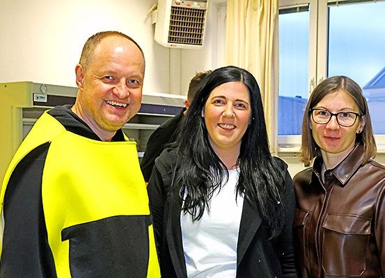 Drei Personen stehen in einem Raum und lächeln für ein Foto. Ein Mann in einem gelben Mantel, eine Frau in einem weißen Shirt und eine weitere Frau in einem braunen Ledermantel. Hinter ihnen zeigt ein Fenster ein Gebäude.