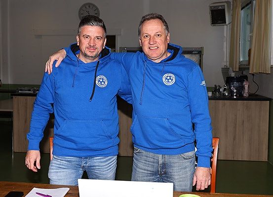 Zwei Männer in blauen Hoodies stehen in einem Raum, lächeln und posieren für ein Foto. Hinter ihnen befindet sich eine Küchenzeile mit einem Tisch, einer Uhr und einem Fenster.