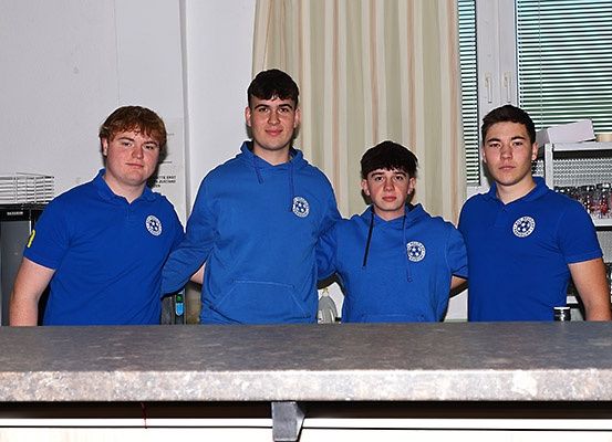 Fünf junge Männer in blauen Uniformen mit einem weißen Logo stehen zusammen hinter einer Theke in einer Küche.